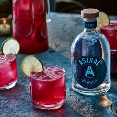 Two magenta drinks fill shorter glasses with wheels of limes on the edge of each glass; a bottle of Astral Tequila is nearby and in the background there's a pitcher of the same drink.