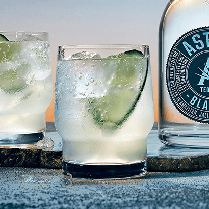 Drink in glass with a large slice of cucumber and ice; bottle of Astral Tequila to the side
