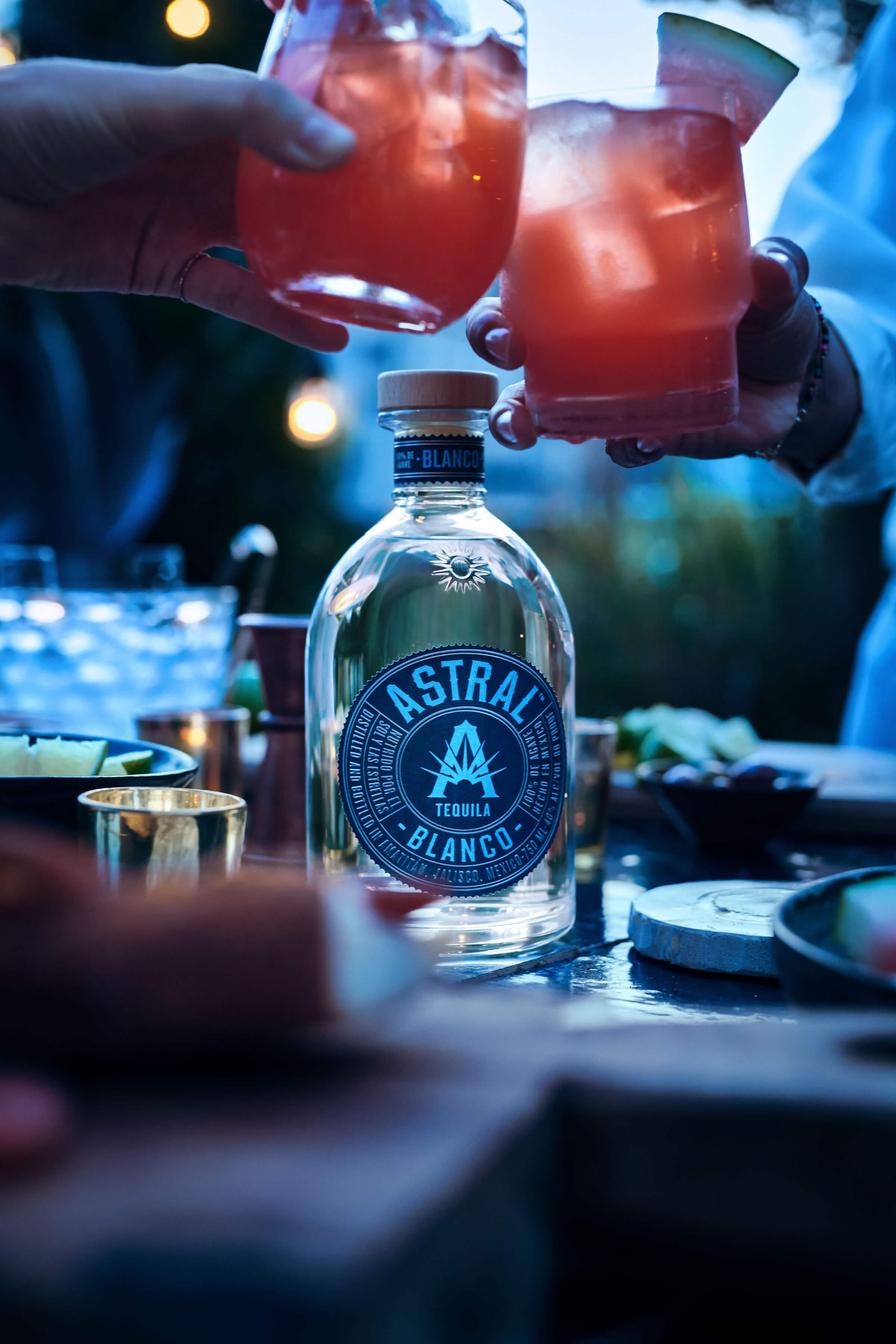 Bottle of Astral Tequila, above hands bring together two glasses in cheers