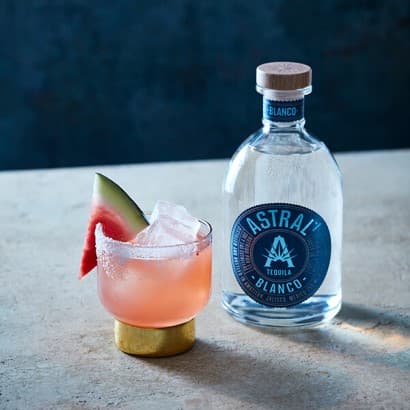 Pink drink in a glass with a gold bottom; ice rests inside and a wedge of watermelon rests on the edge; bottle of Astral Tequila nearby