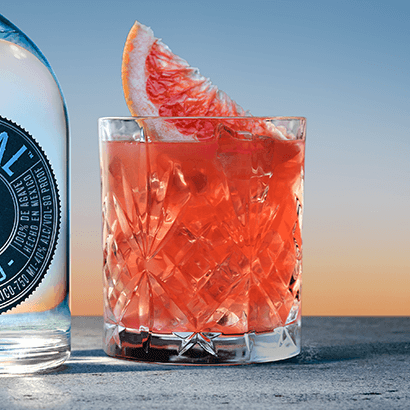 Red-orange drink in a textured glass with a wedge of blood orange resting on top of; bottle of Astral Tequila to the side
