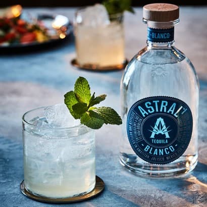 Glass with ice is filled with a pale white cocktail, a sprig of mint decorates the glass; a bottle of Astral Tequila rests nearby.