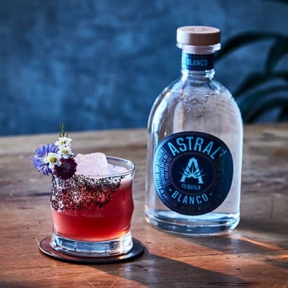 Pink drink in a glass with a hibiscus flower decorating the top; bottle of Astral Tequila nearby