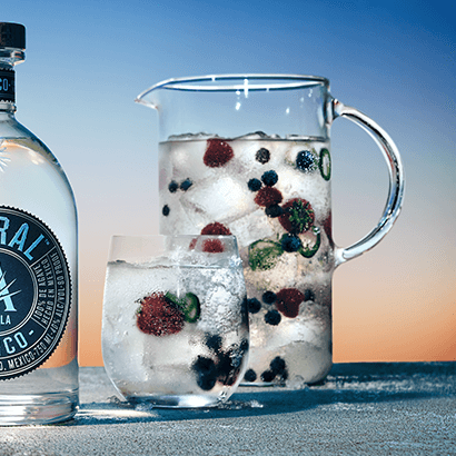 Large pitcher filled with ice, cocktail, and floating berries. Small glass of same drink is in front of the pitcher and a bottle of Astral Tequila is to the side