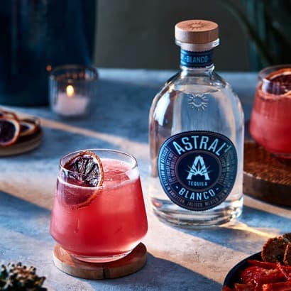Magenta drink in a glass with a slice of blood orange resting on top; bottle of Astral Tequila is next to the glass