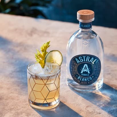 Pale yellow drink in a short glass with a wedge of lime on the edge and a sprig of celery decorating; bottle of Astral Tequila is next to the glass
