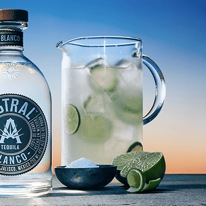Drink in a large pitcher with ice and cucumbers floating inside. Bottle of Astral Tequila is next to the pitcher.