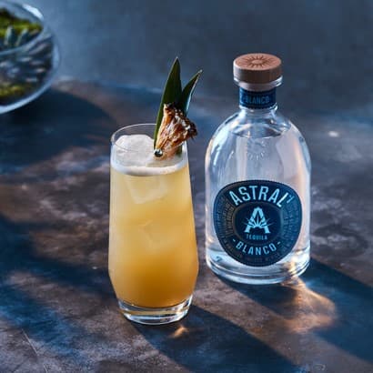 Yellow drink in a tall glass with sage leaves decorating the drink; a bottle of Astral Tequila is next to the glass