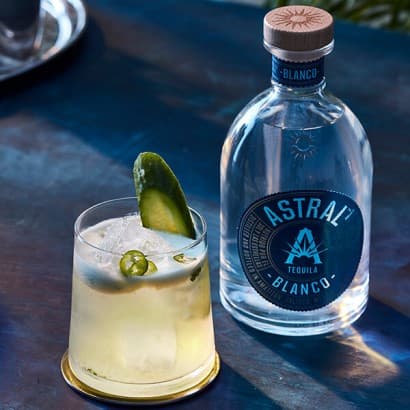 A pale yellow cocktail in a tapered glass with slices of cucumbers and jalapeños and ice; bottle of Astral Tequila is to the side