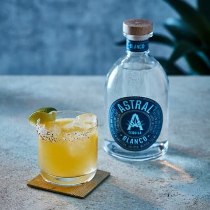 Yellow drink in a glass with a wedge of lime on edge rests on a coaster; bottle of Astral Tequila to the side.