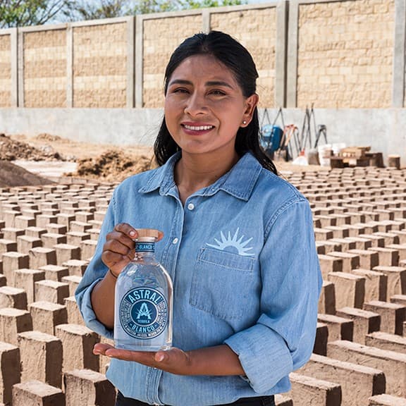 Woman holding a bottle of Astral Tequila, adobe bricks are lined behind her